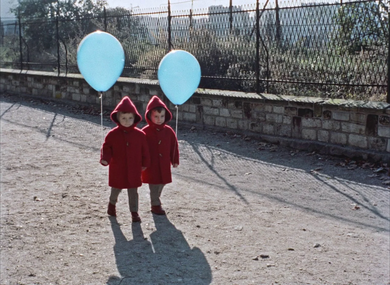 The Red Balloon (1956) movie still: fullbody — A dirt-covered path runs alongside a weathered stone wall and metal wire fence. Two small children…