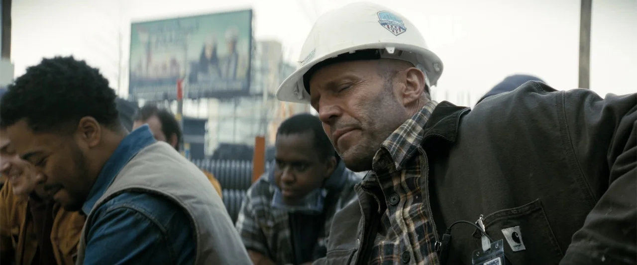 A Working Man (2025) movie still: medium — A construction site features laborers in work gear with a blurred billboard in background. A middle-aged…
