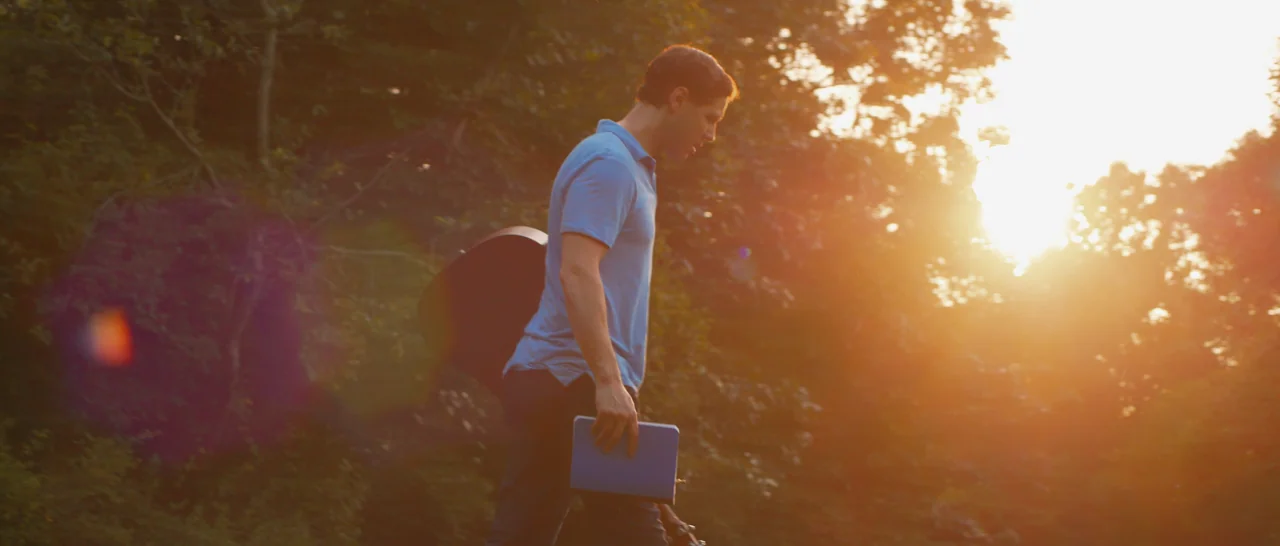 Songs For A Sloth (2021) movie still: fullbody — A man walks through a tree-lined area illuminated by the low afternoon sun. He carries a guitar…