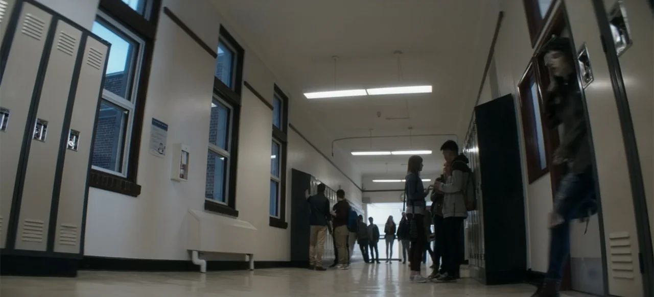 I Still See You (2018) movie still: wide — A high school hallway with rows of metal lockers and windows along the wall. Small groups of students…