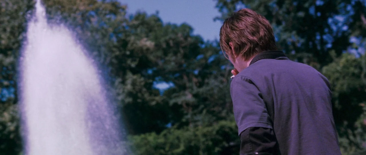 Stay (2005) movie still: medium — A man stands outdoors near a tall water fountain with trees in the background. He holds a cigarette to his mouth…