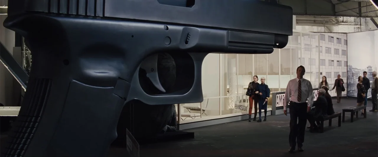 Unfinished Business (2015) movie still: wide — A spacious gallery features a giant model handgun sculpture in the foreground and white partition…