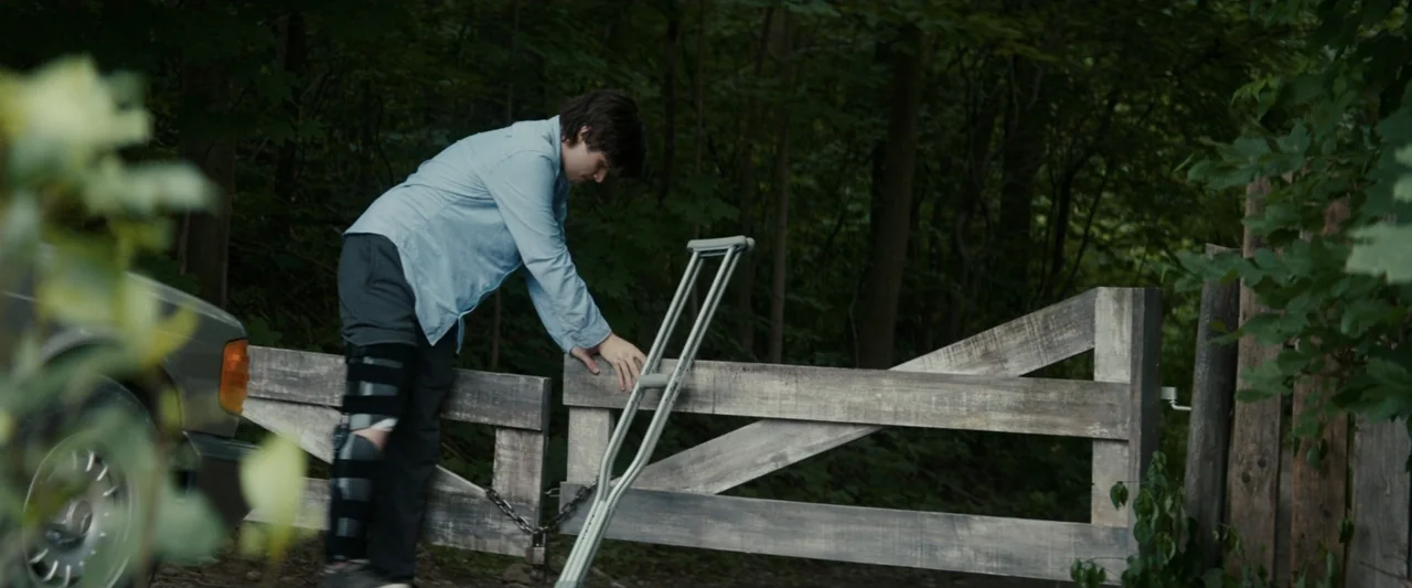 Strange but True (2019) movie still: fullbody — A rural setting features a wooden gate positioned in front of a dense forest. A young man with a leg…
