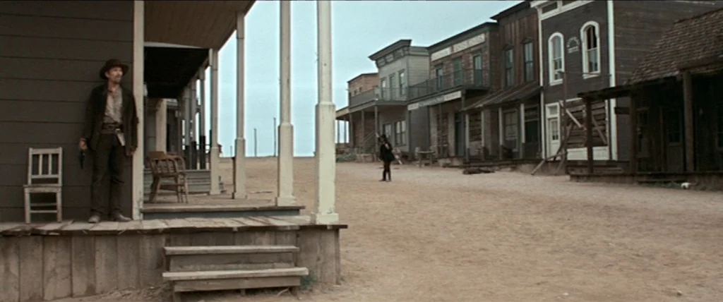 In A Valley of Violence (2016) movie still: wide — A dusty, empty main street of a frontier town features wooden buildings with porches. A man…