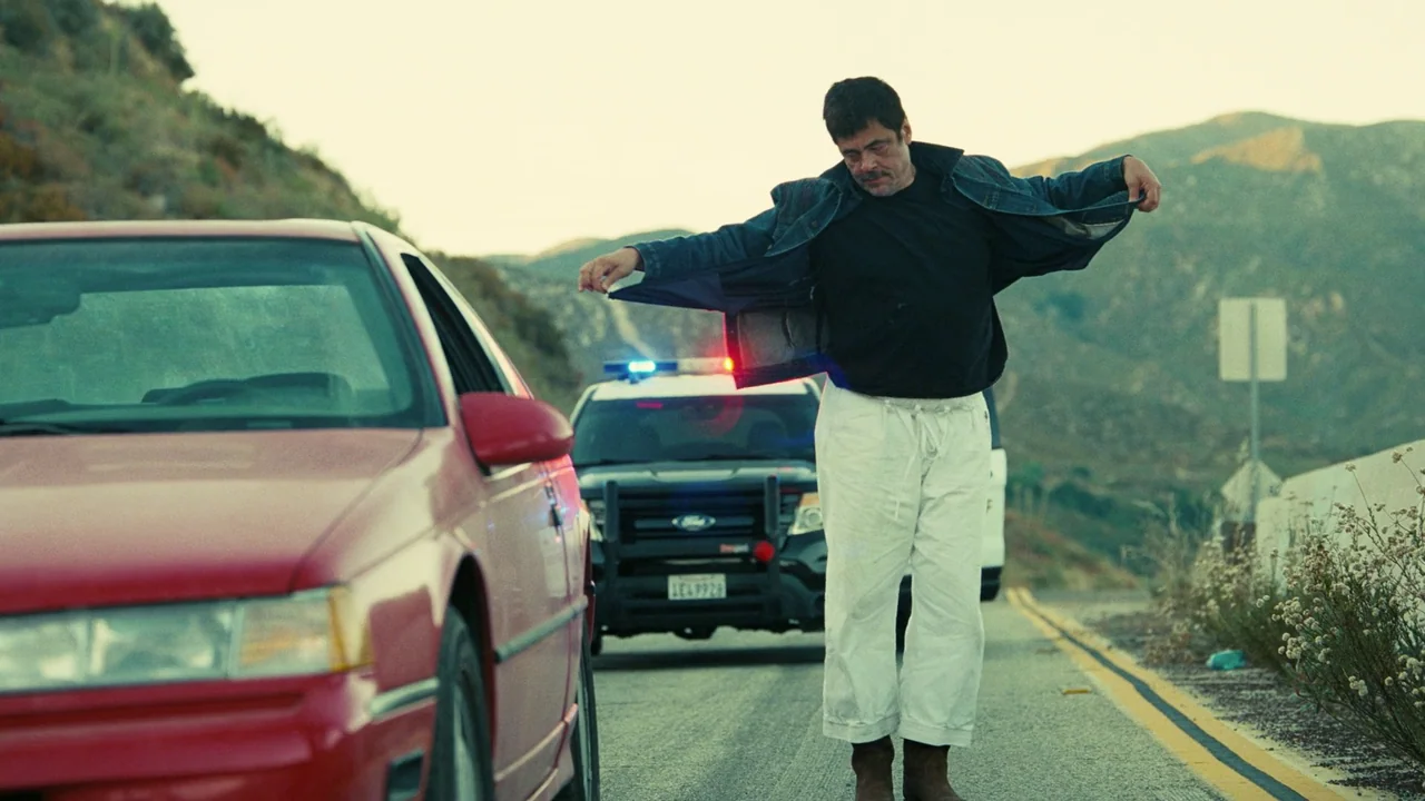 One Battle After Another (2025) movie still: fullbody — A rural highway shoulder with rolling hills and a parked police cruiser in distance. A…