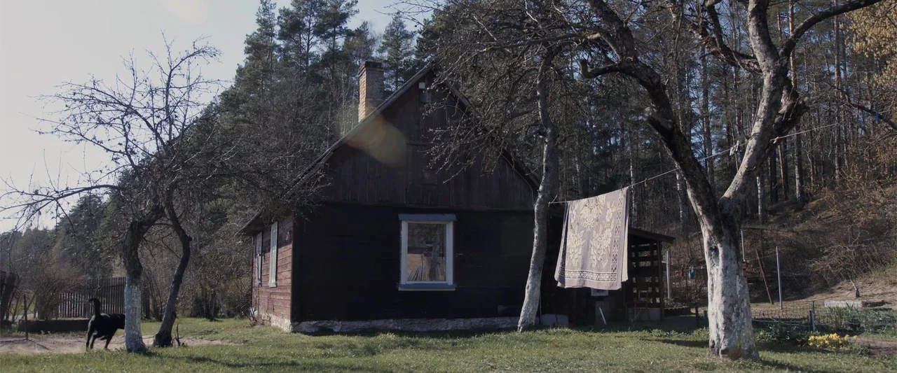 The Good Neighbor (2022) movie still: wide — A wooden cabin stands in a clearing surrounded by a dense pine forest. A patterned rug hangs from a…