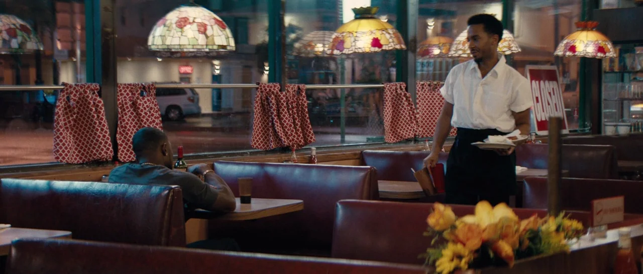 Moonlight (2016) movie still: wide — A diner interior features red vinyl booths and distinctive multi-colored stained glass lamps. A waiter walks…