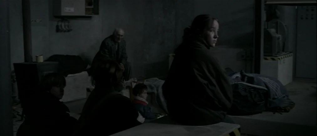 Time Of The Wolf (2003) movie still: medium — A dimly lit concrete basement contains multiple people sitting on raised platforms. They rest amongst…
