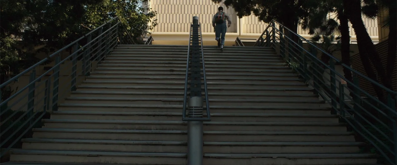 Init!ation (2021) movie still: wide — A wide concrete staircase leads upward toward a building facade with vertical textured panels. A person…