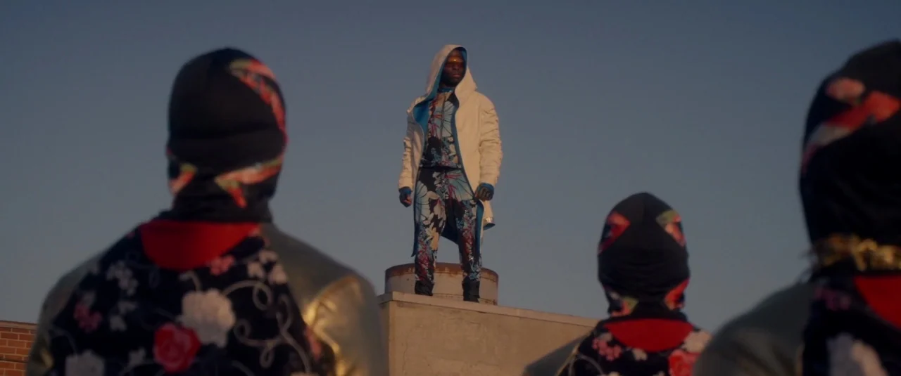 Officer Downe (2016) movie still: fullbody — A man stands atop a vent on a flat concrete building at sunset. Three figures wearing patterned masks…
