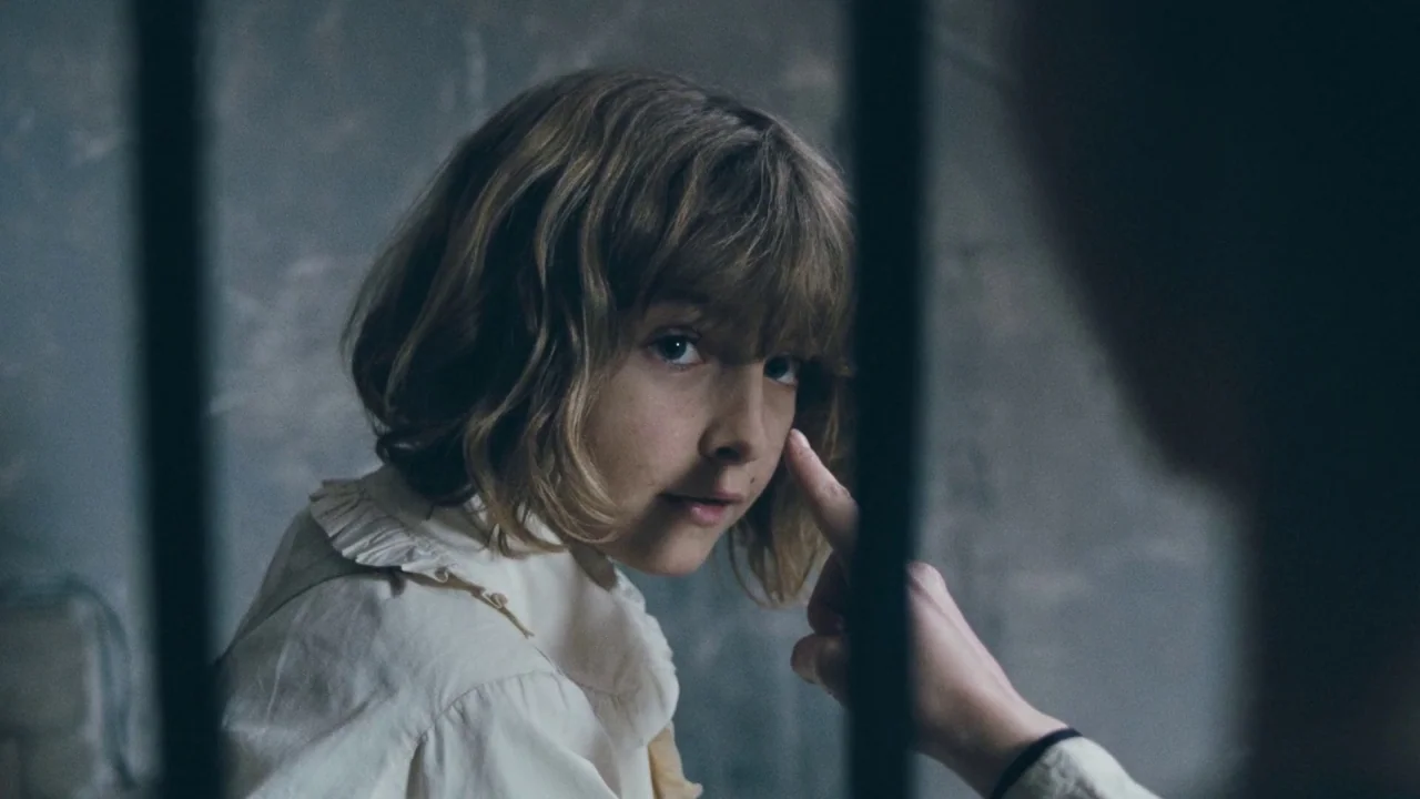 Childhood of a Leader (2015) movie still: closeup — A dimly lit room with stone walls and dark vertical iron bars. A child is being touched on the…