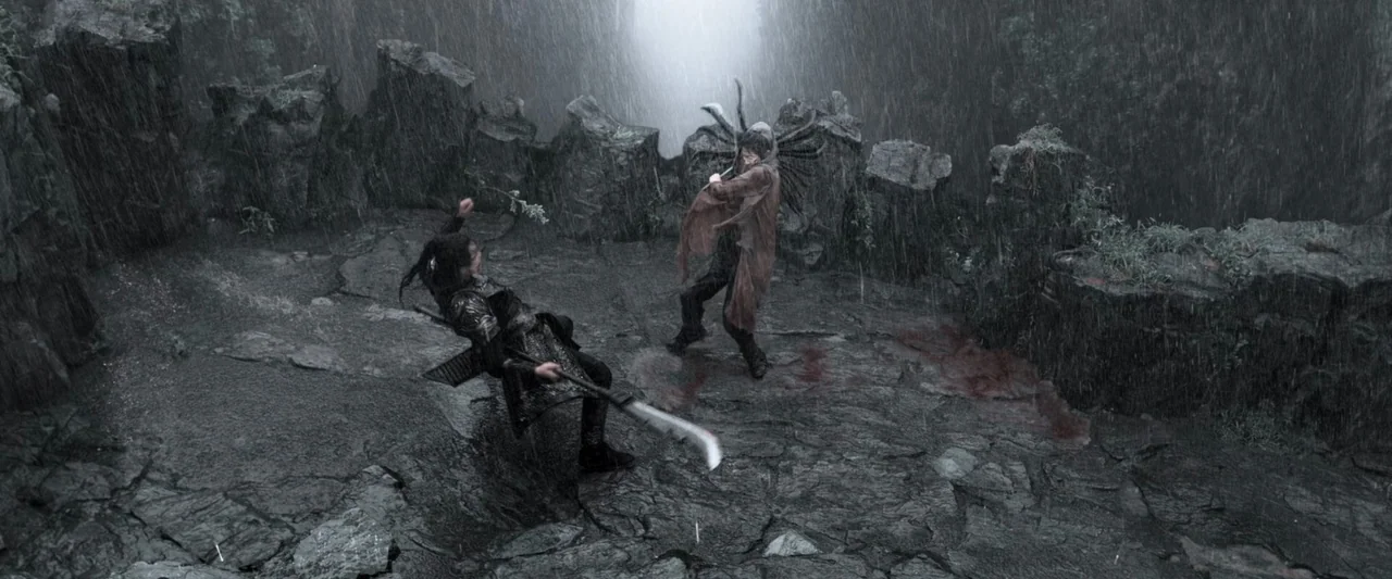 Shadow (2018) movie still: wide — A rain-slicked rocky ledge is surrounded by vertical stone walls under a stormy sky. Two combatants battle with…