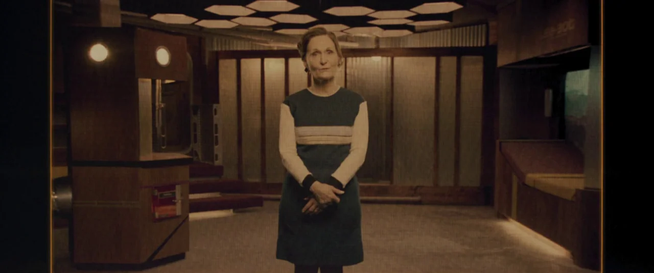 Domain (2017) movie still: fullbody — A 1970s-styled futuristic room with wood paneling and hexagonal ceiling lighting fixtures. An elderly woman…