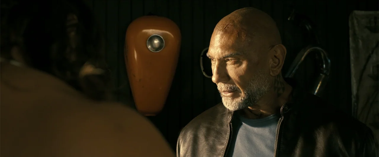 The Wrecking Crew (2026) movie still: medium — A dark workshop space contains an orange motorcycle gas tank hanging on a wall. A man with a grey…