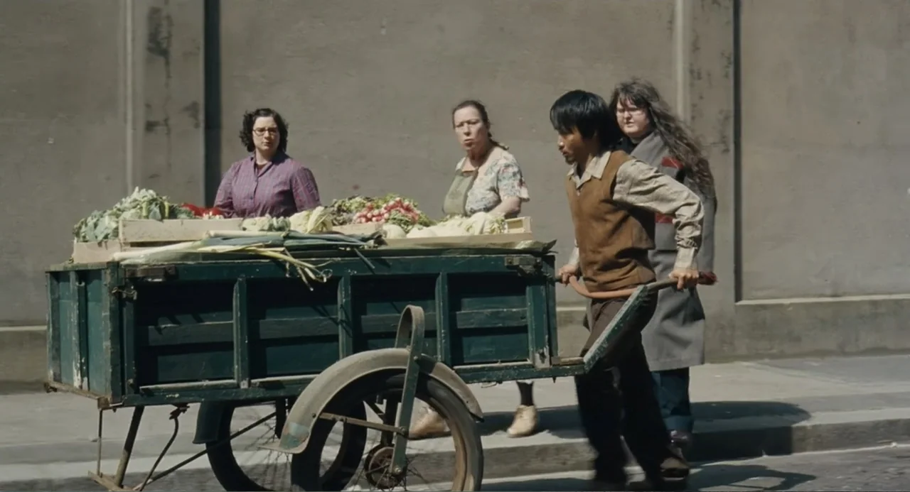 Le Havre (2011) movie still: fullbody — A group of people walk along a sidewalk next to a plain concrete wall. A young man pushes a wooden cart…