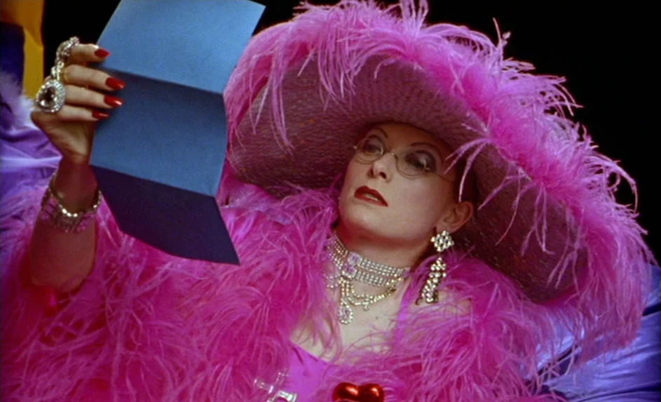 Wittgenstein (1993) movie still: closeup — A woman wears an elaborate pink feathered ensemble and jewelry while lying down. She holds a rectangular…