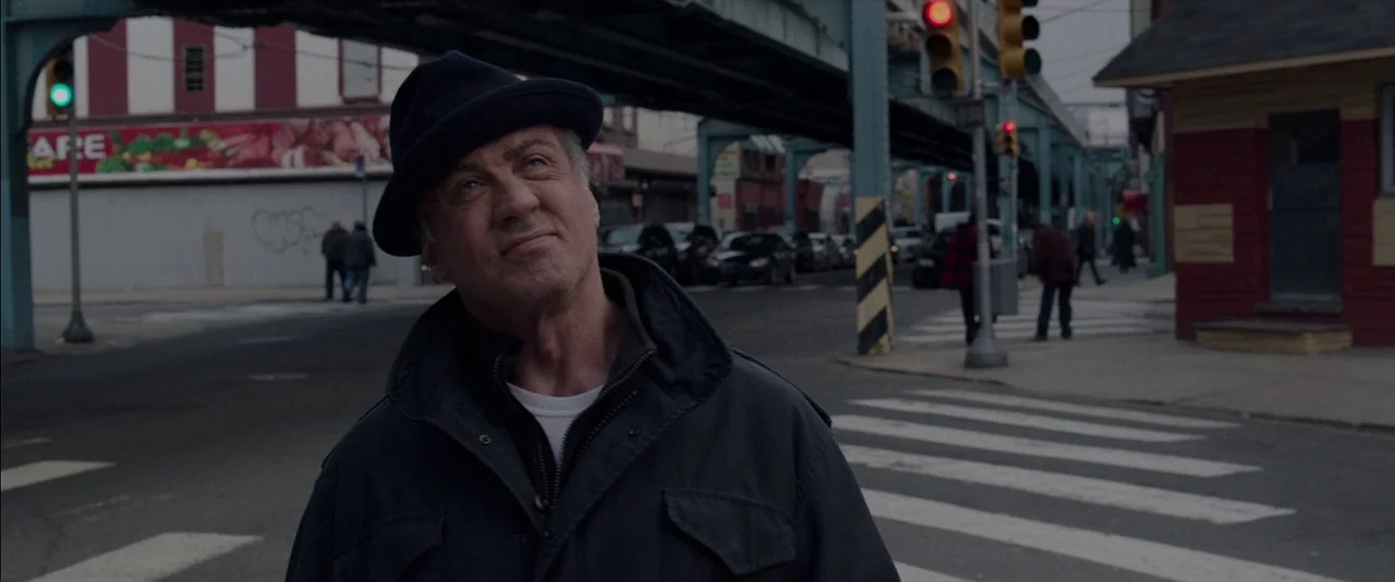 Creed (2015) movie still: medium — A city intersection sits beneath elevated train tracks with a crosswalk in the foreground. An older man stands in…
