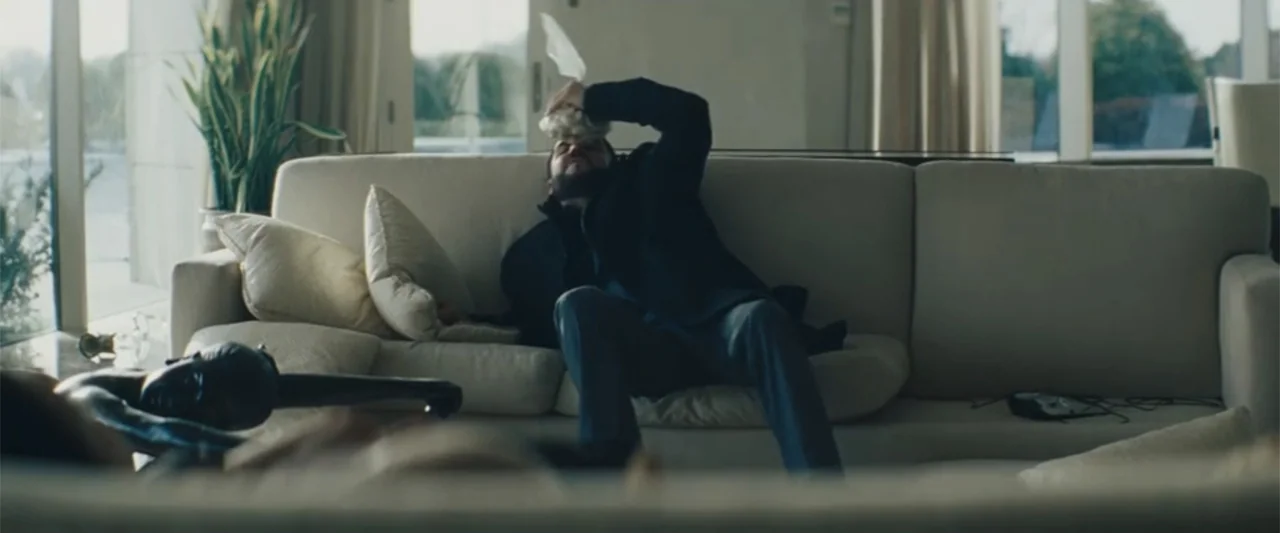 Anora (2024) movie still: medium — A contemporary minimalist living room with white sofas and large floor-to-ceiling windows. A man sits on the sofa…