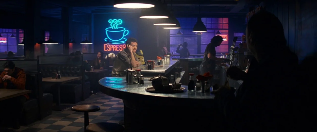 Pokémon Detective Pikachu movie still: wide — A dimly lit modern diner features checkerboard floors and blue neon signage on walls. A young man sits…
