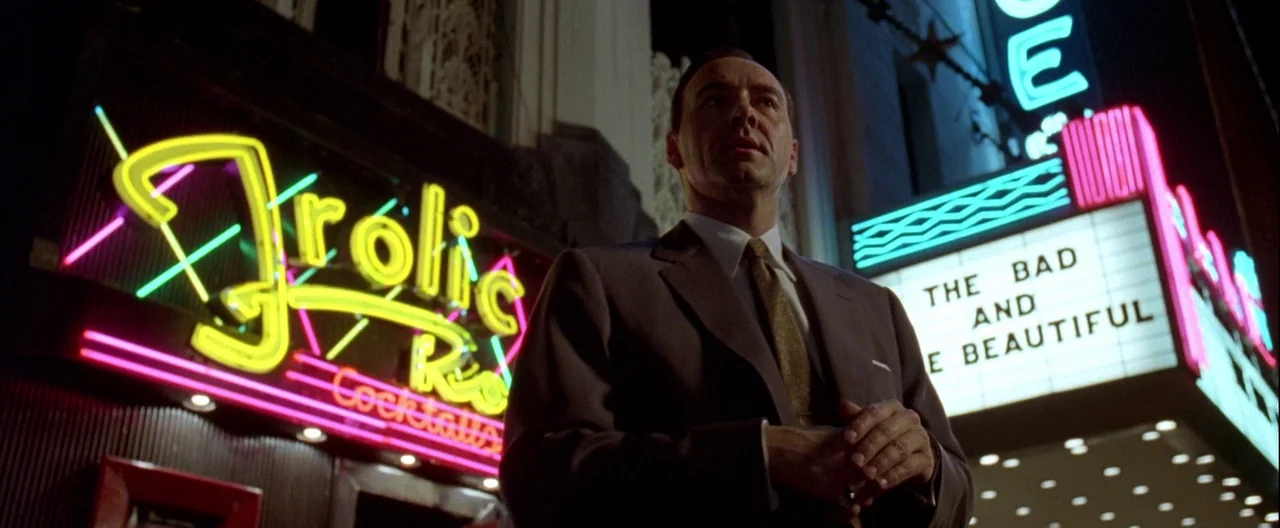 L.A. Confidential (1997) movie still: medium — A man stands outside a vintage movie theater with bright neon signage at night. The marquee displays…