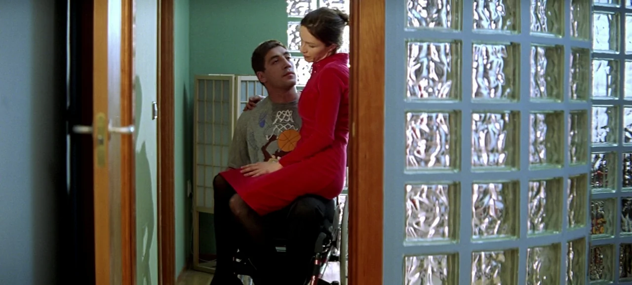 Live Flesh (1997) movie still: medium — A modern room features a grid of glass blocks against the wall. A woman in a red dress sits on the lap of a…