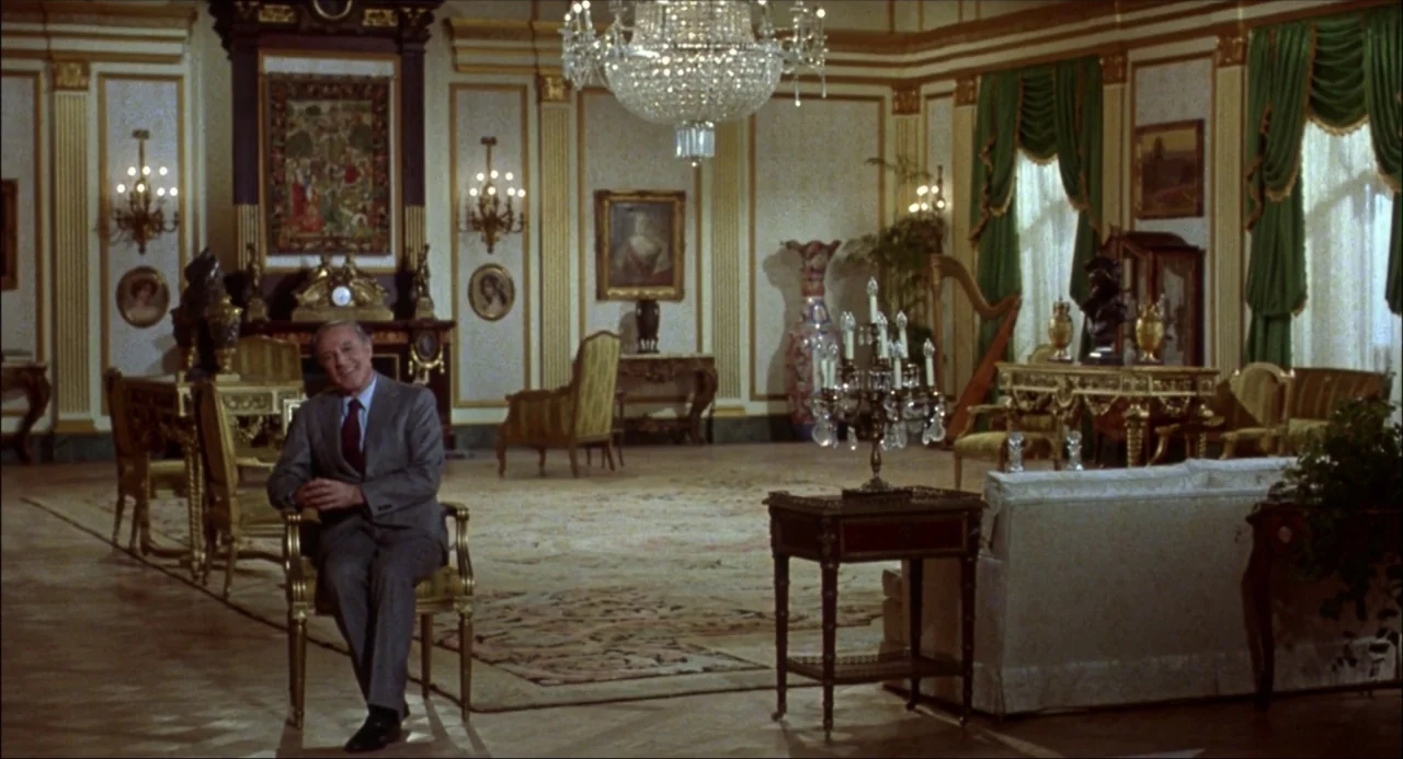 Xanadu (1980) movie still: fullbody — A spacious, ornate hall featuring gilded wall moldings and polished parquet flooring. An elderly man sits on a…