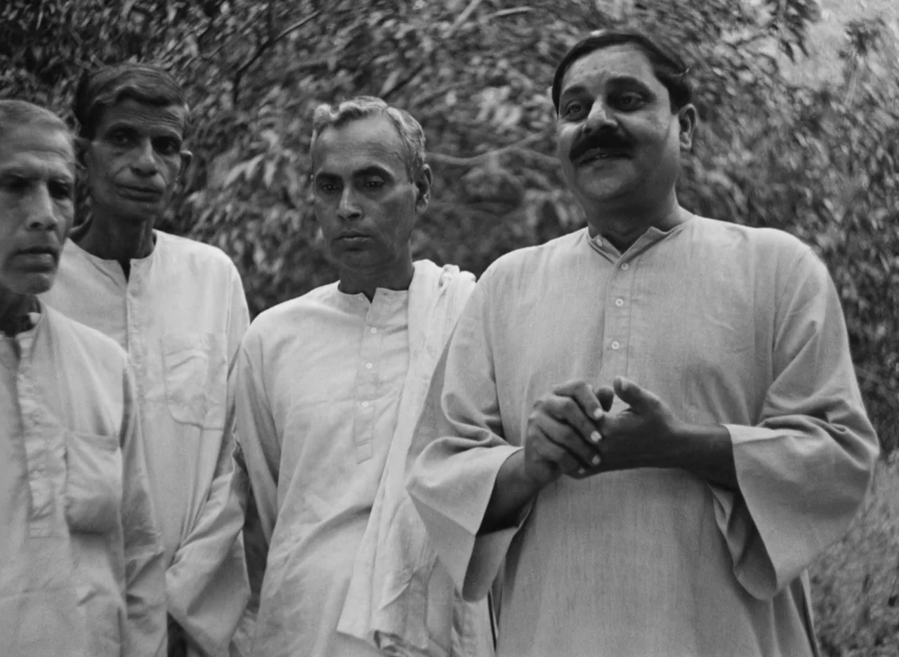Apur Sansar (1959) movie still: medium — Four men stand together in a wooded area among dense, out-of-focus foliage. They wear simple traditional…
