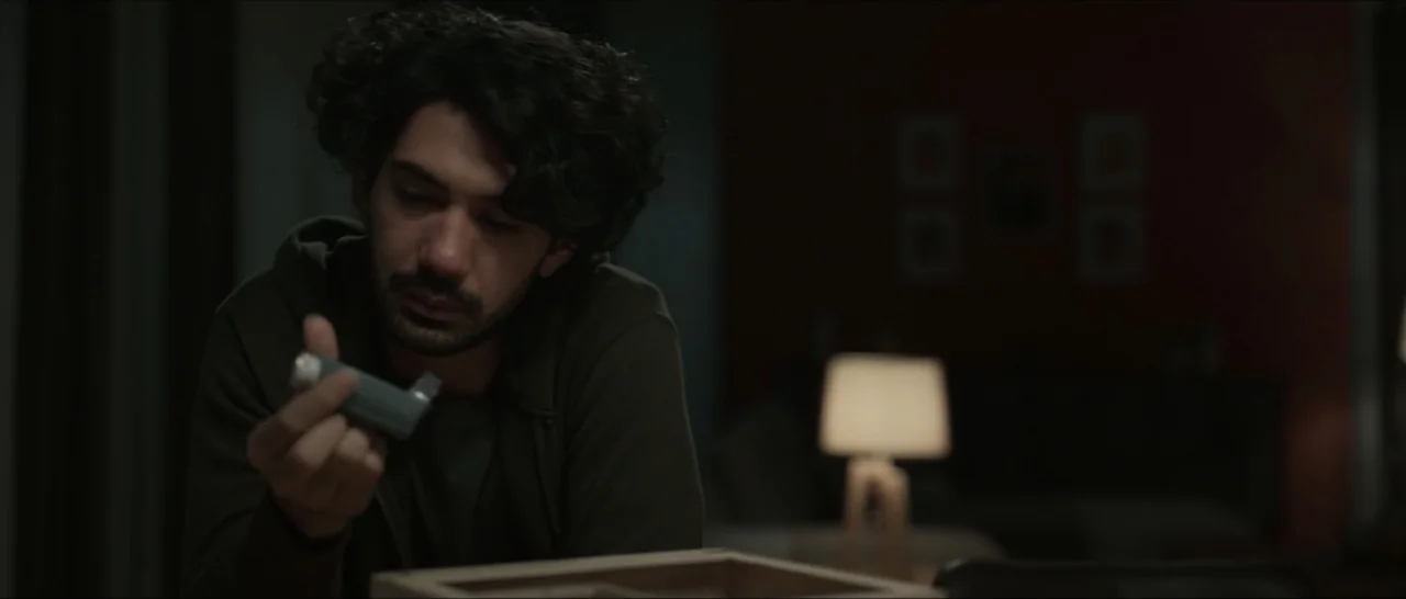 Innocent Vengeance (2023) movie still: medium — A dimly lit interior space with a table and a blurred lamp in the background. A young man with dark…