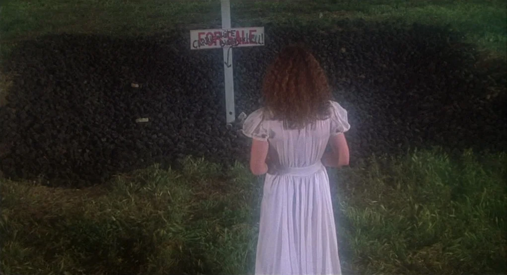 Carrie (1976) movie still: fullbody — A night scene shows a dark rectangular pit in a grassy field at night. A white woman in a white dress stands…