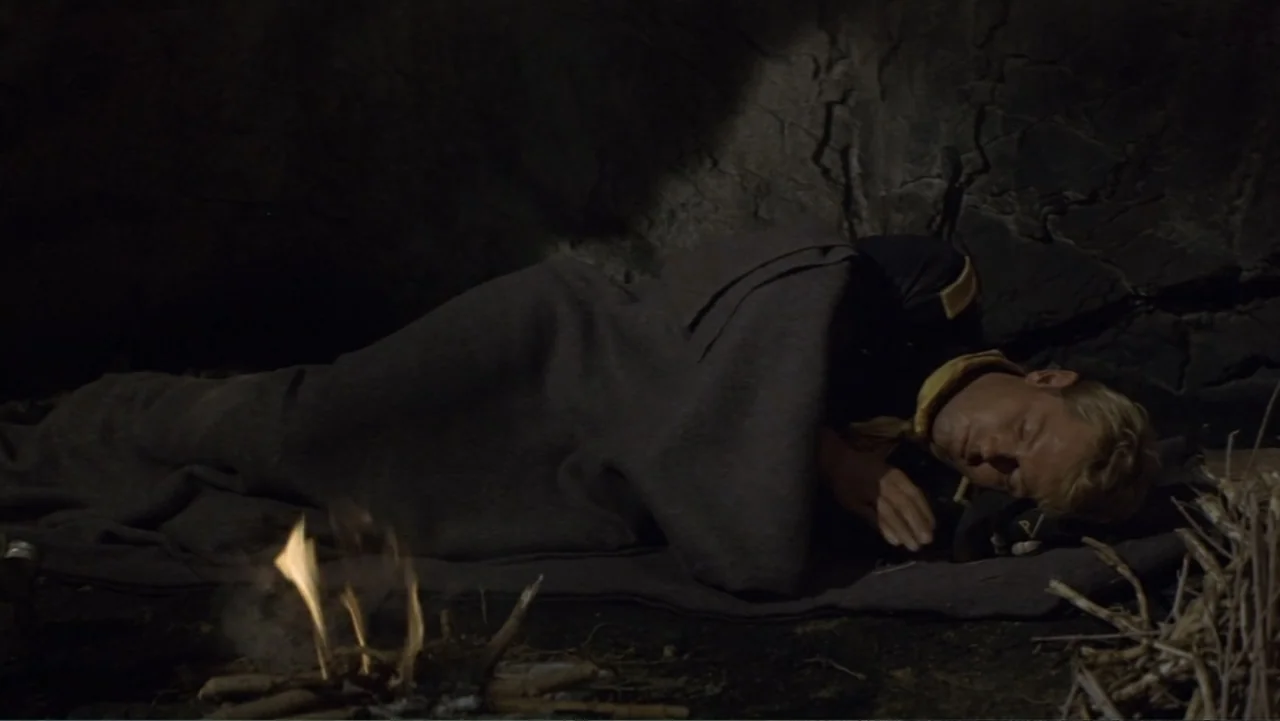 A Distant Trumpet (1964) movie still: medium — A dark, rocky cave interior with a small crackling campfire on the ground. A young man in a military…