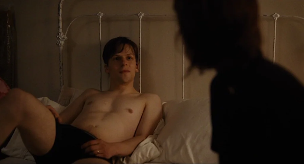 Louder Than Bombs (2015) movie still: medium — A dimly lit room contains a white iron-framed bed against a plain wall. A man sits shirtless in the…