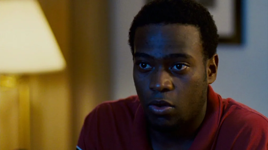 Sugar (2008) movie still: closeup — A dimly lit room contains a man seated near a soft-focus warm lamp. The man looks toward the left of the frame…