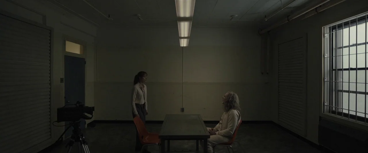 Longlegs (2024) movie still: wide — An austere room with beige walls and a metal table in the center. A woman stands opposite a man sitting at the…