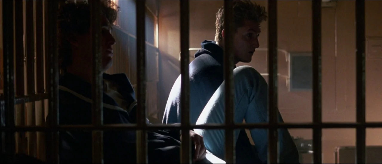 At Close Range (1986) movie still: medium — A cramped room with metal bars blocking the view of two men inside. The pair sits on a bench while…