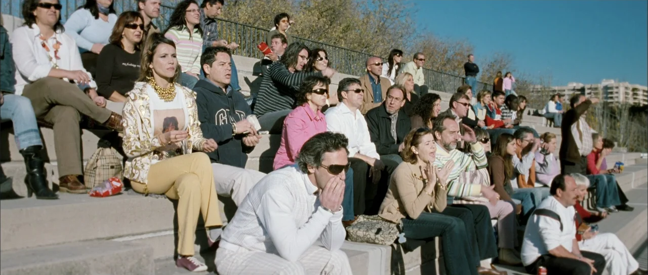 Rivals (1972) movie still: wide — A crowd sits on tiered concrete bleachers outdoors on a sunny day. Many people watch an event unfolding off-screen…