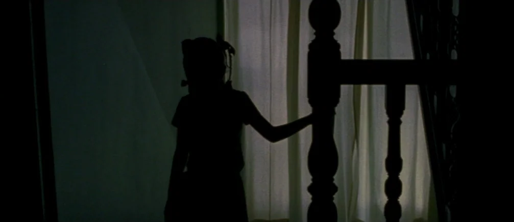 Innocence (2005) movie still: medium — A dark, silhouetted child stands beside a wooden staircase post in front of curtains. The scene is framed to…