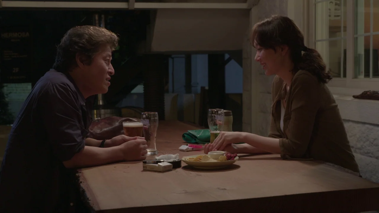 Yourself and Yours (2016) movie still: medium — An outdoor patio at night features a large live-edge wooden table with drinks. A man and woman sit…