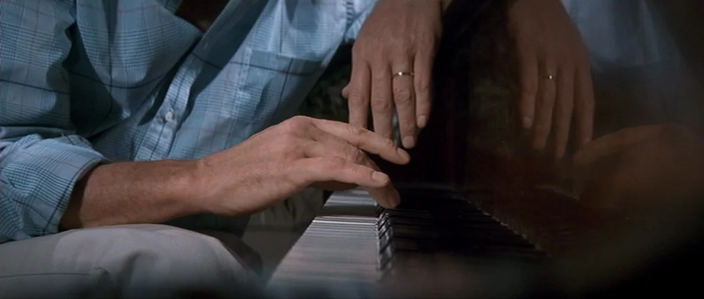 Cape Fear (1991) movie still: closeup — A person in a blue plaid shirt sits before the keys of a piano. Their hands rest on the keys with a gold…