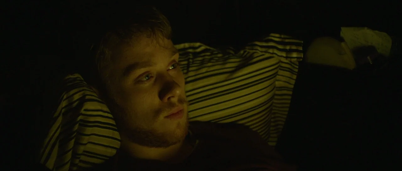 Green Room (2015) movie still: closeup — A man lies on a striped pillow in a dark, unlit room. The subject gazes ahead, illuminated by a warm,…
