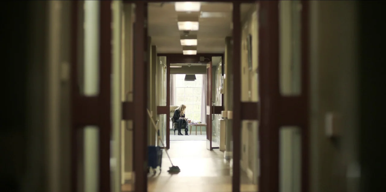 Help (2021) movie still: wide — A long institutional corridor with glass doors leads to a seating area with a window. A woman sits in a solitary…