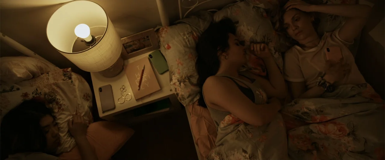 Init!ation (2021) movie still: medium — A cluttered bedside table sits between two beds in a softly illuminated room. Three young women lounge or…