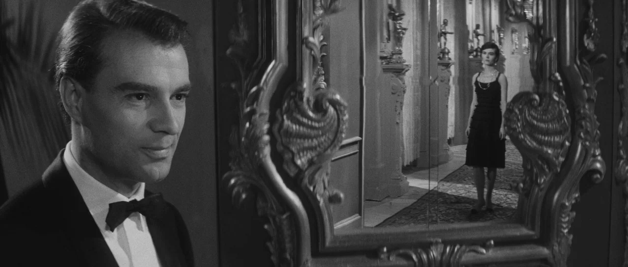 Last Year at Marienbad (1961) movie still: medium — A man in a tuxedo stands in the foreground watching a woman in a mirror. The ornate, carved…