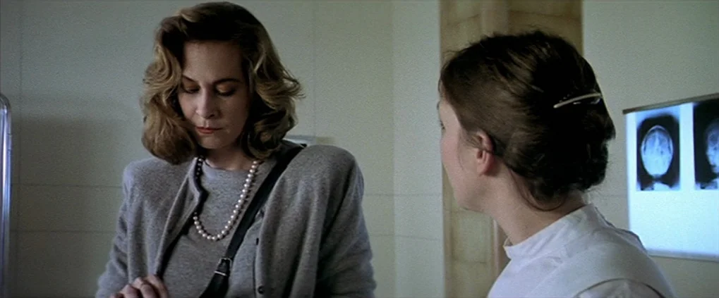 Anguish (1987) movie still: medium — Two women stand in a clinical, white-walled room with diagnostic imagery mounted behind them. The woman in the…