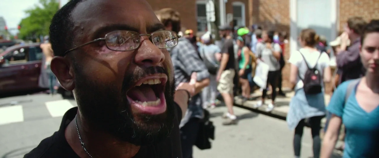BlacKkKlansman (2018) movie still: closeup — A man stands on a busy urban sidewalk filled with a crowd of pedestrians. The man shouts loudly with…