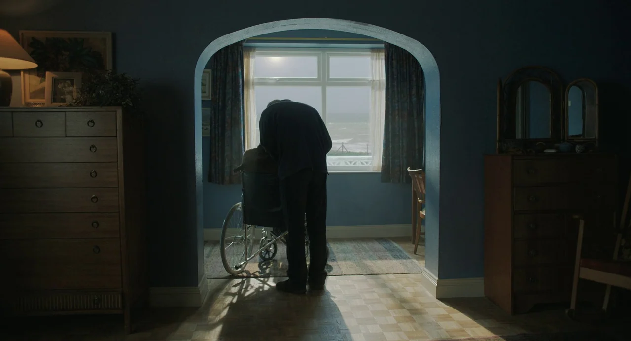 My Policeman (2022) movie still: medium — A furnished interior with blue walls features an arched entryway overlooking the seaside coast. An elderly…