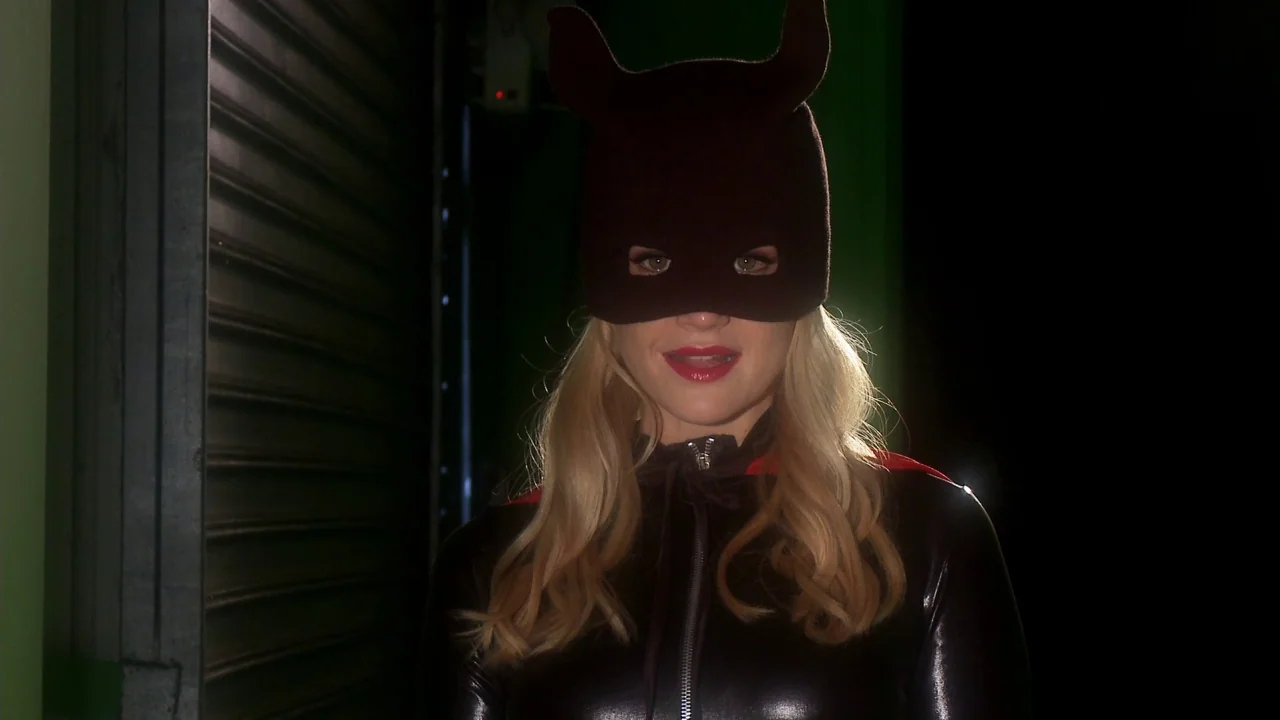 Electra Luxx (2010) movie still: medium — A dark space with a green painted wall and a metallic roll-up door. A blonde woman wearing a black latex…