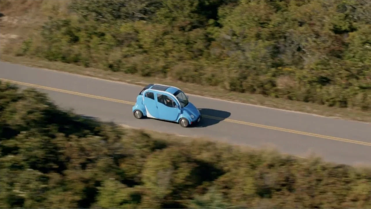 Clear History (2013) movie still: wide — An asphalt road cuts through dense green coastal vegetation on a sunny day. A small blue commuter car…