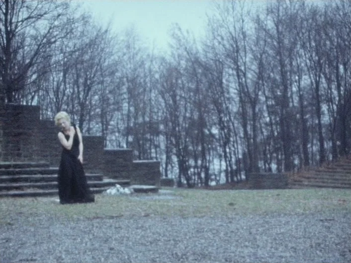 Eika Katappa (1969) movie still: fullbody — A stone stepped memorial sits amidst a grove of bare trees under an overcast sky. A woman in a black…