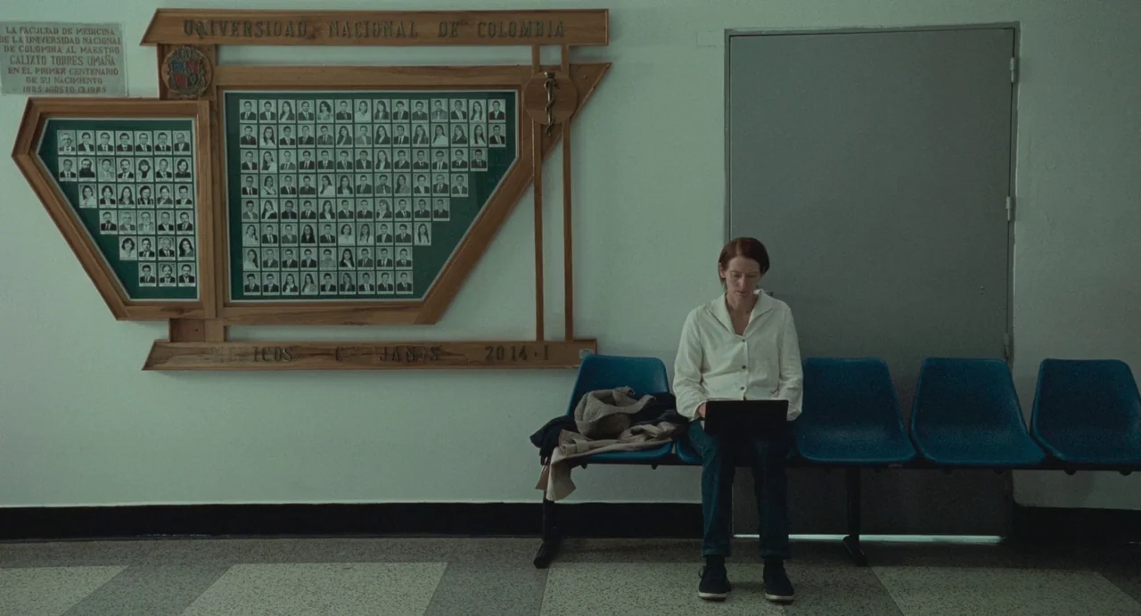 Memoria (2021) movie still: wide — A sterile medical waiting area features a large commemorative plaque with many small portrait photos. A woman…