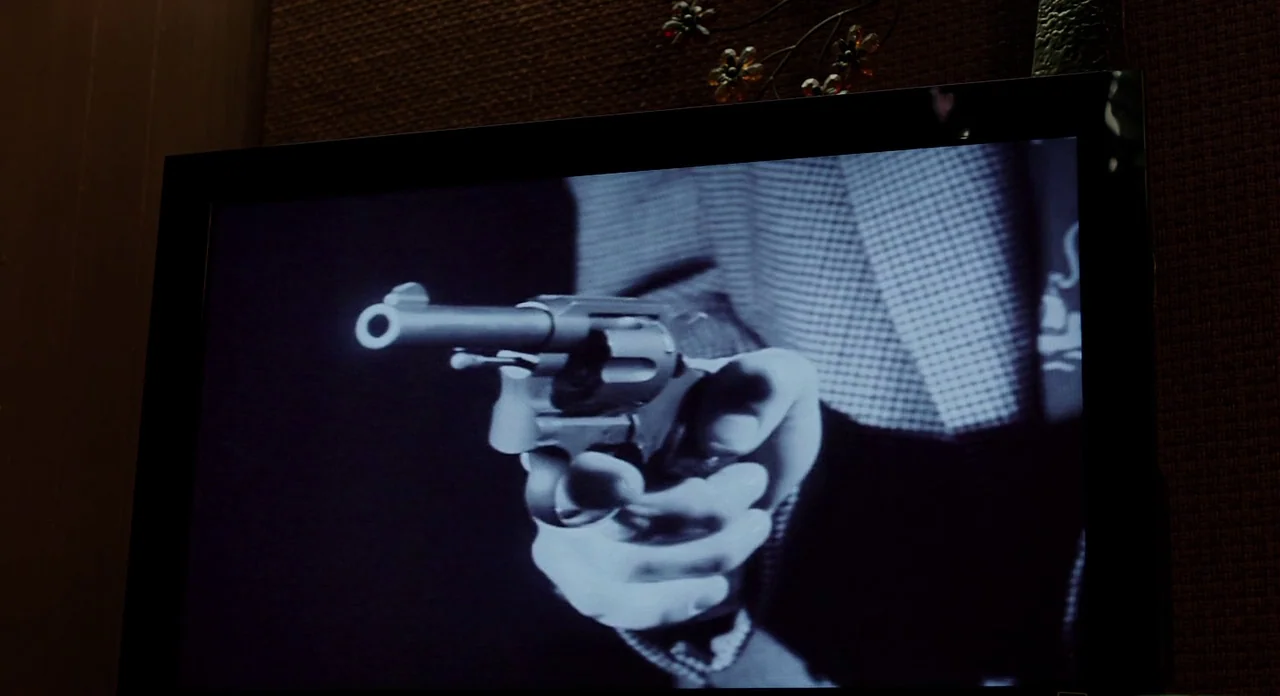 Killer Joe (2011) movie still: closeup — A dark television screen displays a high-contrast black and white film frame. A man's hand holds a revolver…